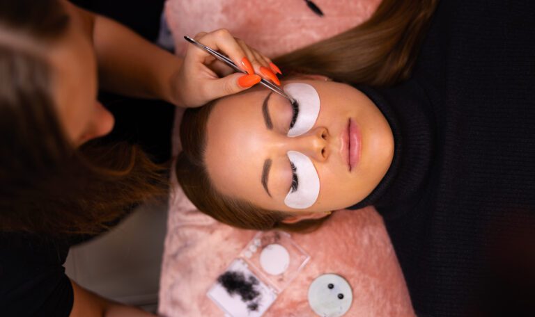 Overhead view of female client going through eyelash extension procedure with lash artist using tweezers
