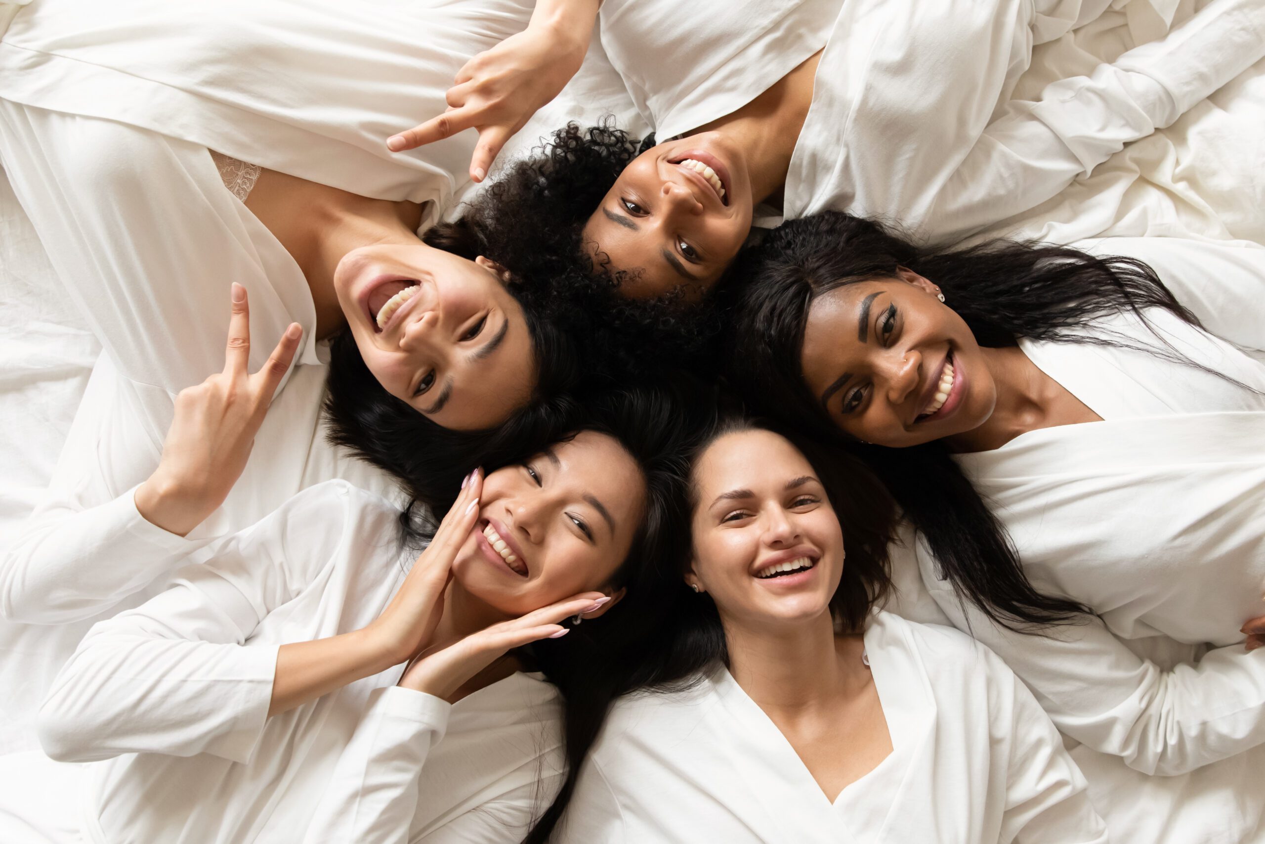Top view five diverse women in white bathrobes lying in bed smile look at camera feels happy after lash extensions