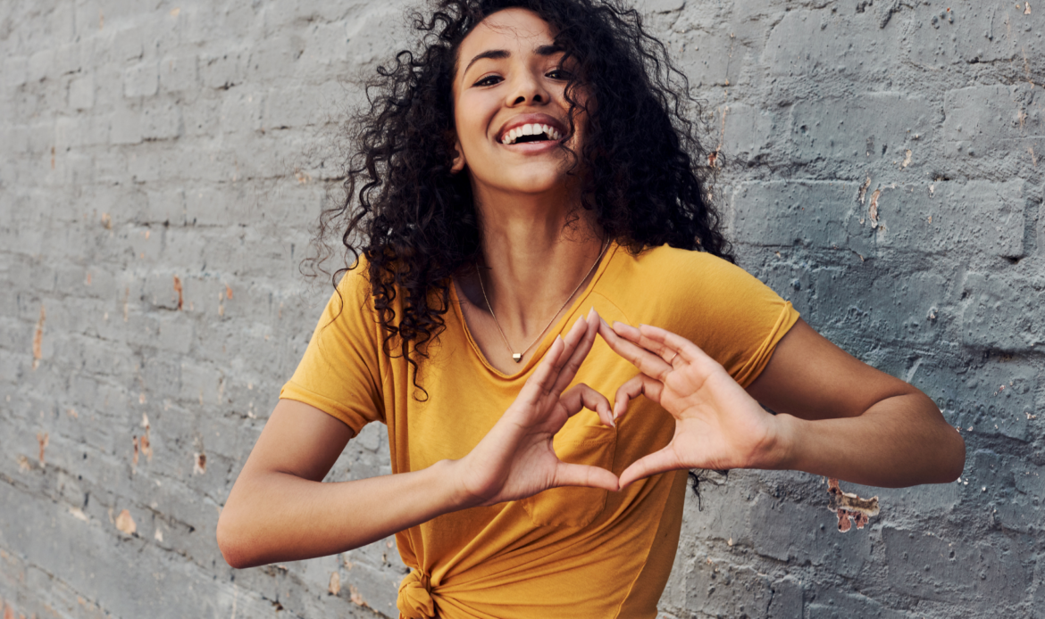 African American young woman in a yellow shirt making a heart with her hands smiling